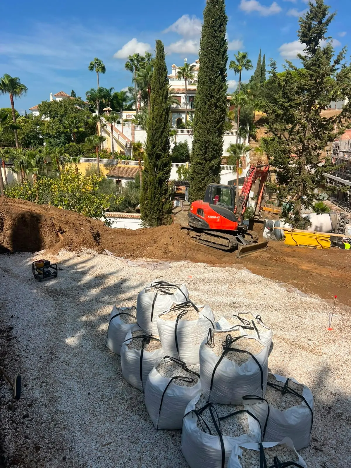 Excavator working the soil mound at El Rosario, with bulk bags of aggregate staged on the site